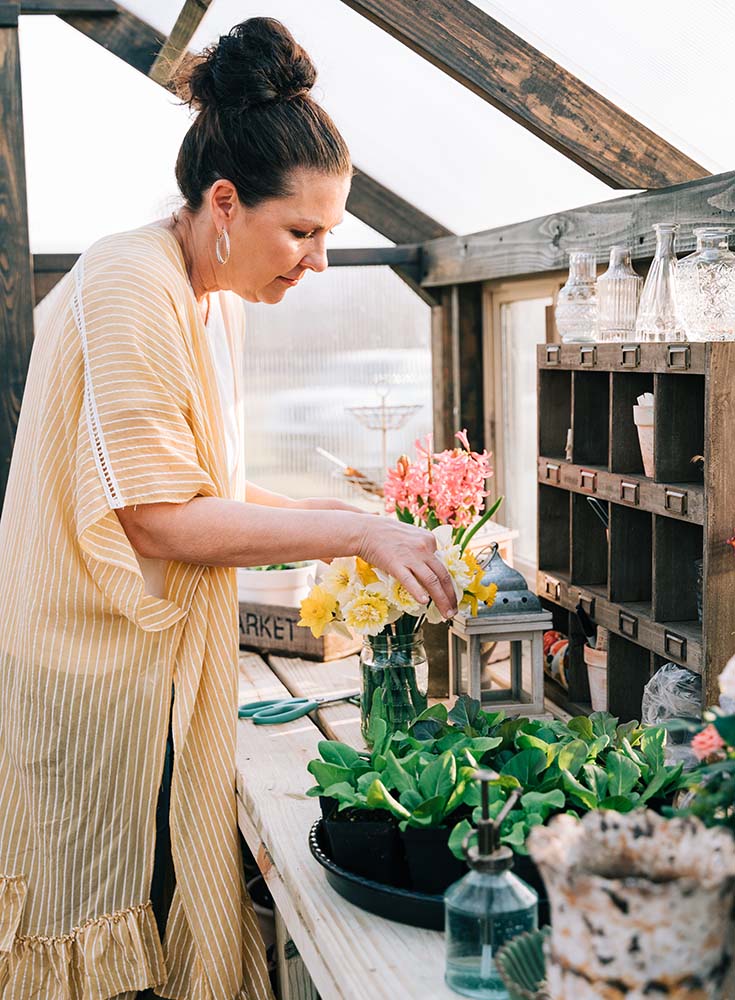 Woman putting yellow flowers into a vase. She is standing inside a greenhouse.