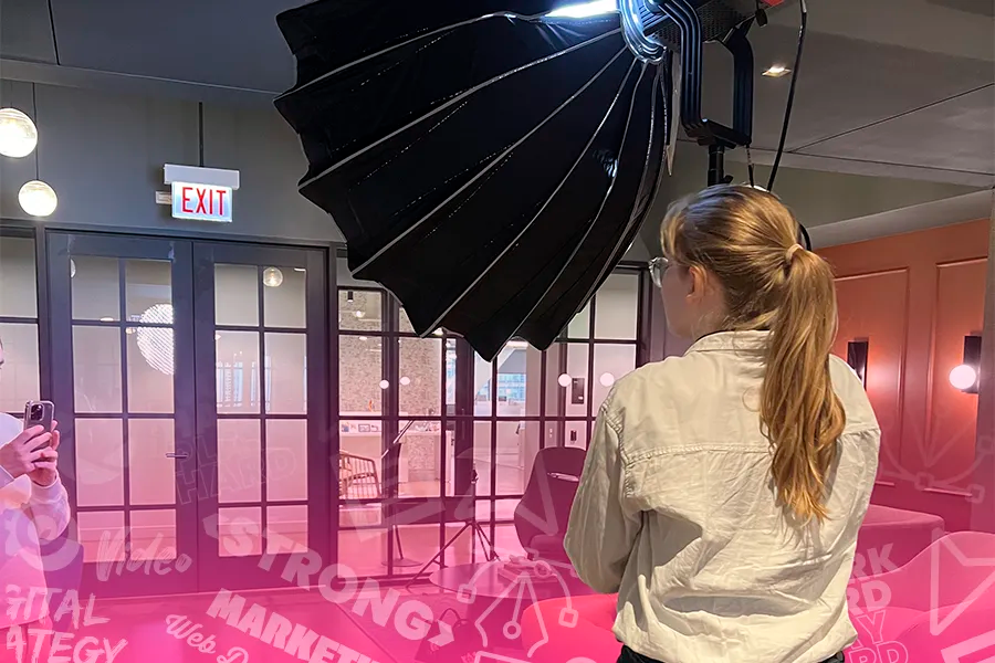 A woman with long hair faces away from the camera to set up lighting for a video shoot in a small office room. Words and phrases like "marketing", "video", "digital strategy", "", and "web design" are lightly laid over the image near the bottom.