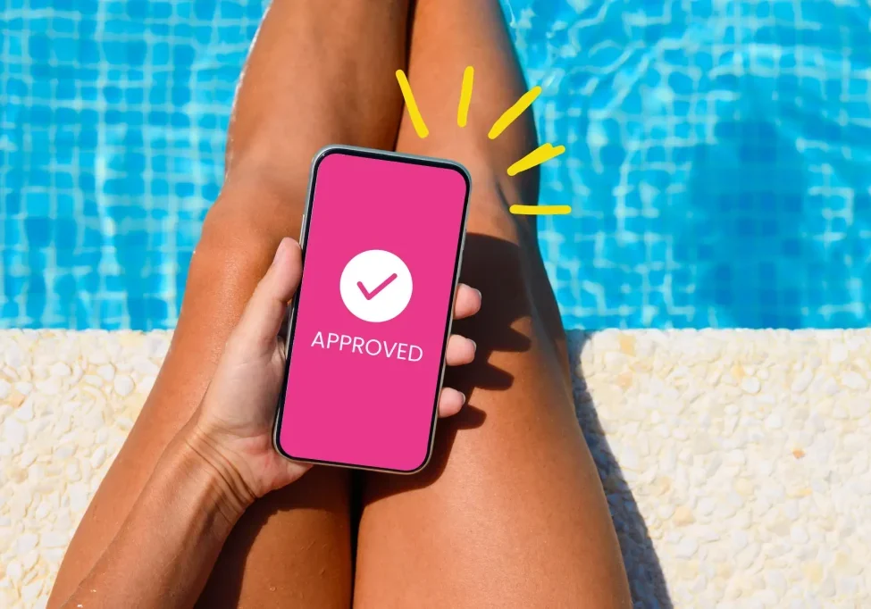 A person's hand holding a smartphone showing a pink "APPROVED" notification screen with a white checkmark. The background shows the person lounging by a bright blue swimming pool on a sunny day.
