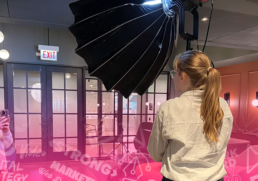 A woman with long hair faces away from the camera to set up lighting for a video shoot in a small office room. Words and phrases like "marketing", "video", "digital strategy", "", and "web design" are lightly laid over the image near the bottom.